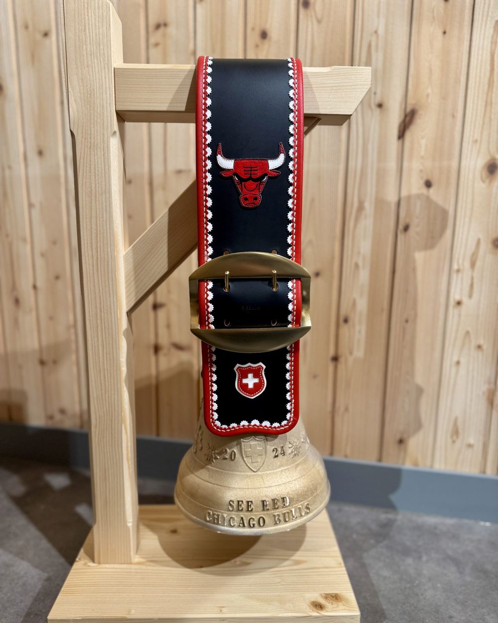 Une cloche et sa courroie brodée d'un taureau rouge et d'une croix suisse, sur un support en bois